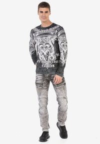 Langärmliges schwarz-weißes Grafik-T-Shirt mit detaillierten Designs. Hellgraue, verwaschene Jeans mit Nieten und Reißverschlussakzenten, kombiniert mit schwarzen Sneakern.