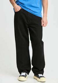 Osoba w luźnych czarnych spodniach, czarno-białych trampkach i niebieskiej koszuli, z jedną ręką w kieszeni na jednolitym tle.