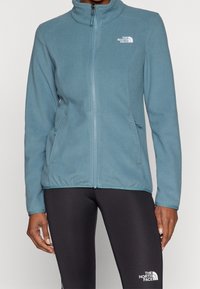 Fleece-takki vaaleansinisellä värillä, jossa on korkea kaulus, etuvetoketju ja sivutaskut. Vasemmassa rinnassa valkoinen logo.