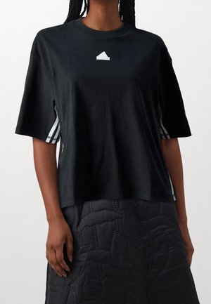 Kobieta w czarnej koszulce Adidas z białym logo i paskami po bokach, zestawiona z pikowaną czarną spódnicą.
