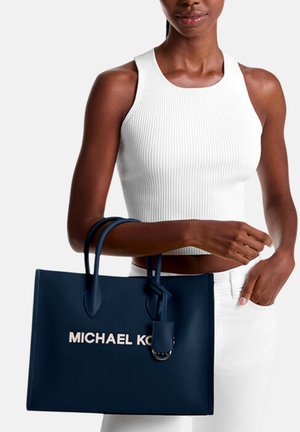 Žena u bijelom rebrastom topu i bijelim hlačama drži veliku tamnoplavu Michael Kors torbu za ručke.
