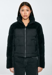 Černá péřovka s huňatým límcem a rukávy, vybavená zipem v přední části a bočními kapsami. Hladká a strukturovaná tkanina se kontrastuje.