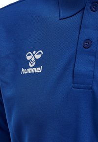 Blå poloshirt lavet af tekstureret stof med en tre-knaps placket. Brandlogoet "hummel" er trykt i hvid på venstre brystområde.