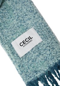 Lys blå tekstureret tørklæde med frynser, med en hvid label der står "CE CIL ACCESSORIES." Blødt materiale og en gradient effekt.