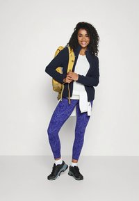 Zestaw odzieży sportowej składający się z granatowej kurtki na zamek, białej koszulki, fioletowych legginsów z wzorem oraz czarnych butów sportowych, uzupełniony żółtym plecakiem.