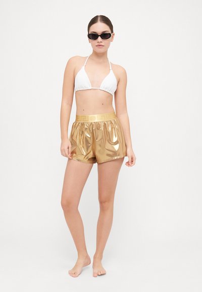 Haut de bikini triangulaire blanc associé à un short doré brillant avec une taille froncée et un motif en relief sur la ceinture.