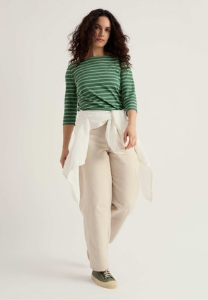 Mujer con cabello rizado que lleva una blusa verde con rayas, pantalones beige, camisa blanca atada a la cintura y zapatillas verdes, de pie frente a un fondo liso.