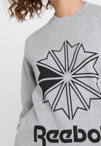 Szary sweter z okrągłym dekoltem, z dużą czarną graficzną gwiazdą w kształcie geometrycznym i napisem "Reebok" w dużych czarnych literach na dole.