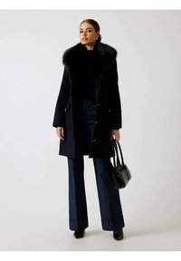 Abrigo de lana negro con cuello de piel sintética, diseño cruzado, botones dorados, combinado con pantalones de mezclilla oscuros de corte acampanado y un pequeño bolso negro.