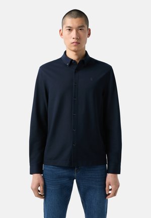 Dunkelblauses Langarmhemd mit Button-Down-Kragen, aus weichem Stoff gefertigt, mit schmaler Passform und dezentem Logo auf der Brust. Kombiniert mit blauen Jeans.