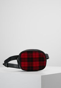 Sac en tissu écossais rouge et noir avec une forme rectangulaire arrondie et une bandoulière en cuir noir. Comprend une fermeture éclair et une texture lisse.