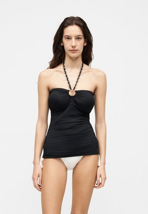 Naine kannab musta rihmadeta topi koos kett-kaelusega ja valgeid bikiinipõhja, seisab tavalise valge tausta ees.