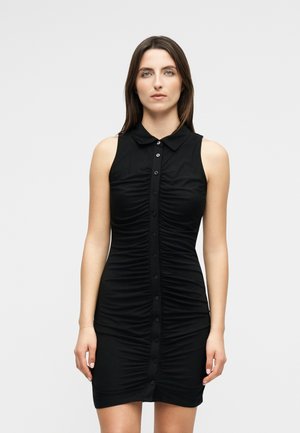 Γυναίκα με ίσια καστανά μαλλιά, φοράει αμάνικο μαύρο φόρεμα με κουμπιά και ρελιαστά πιέτες, σε ανοιχτό γκρι φόντο.