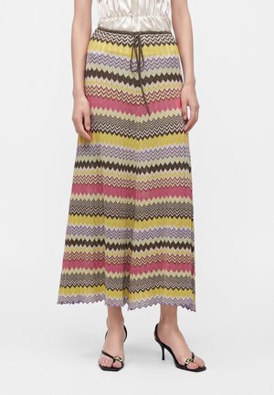 Femme aux longs cheveux raides portant un haut blanc sans manches et un pantalon ample à motifs zigzag colorés avec des talons ouverts noirs, debout devant un fond uni.