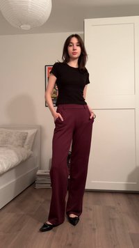 T-shirt noir cintré, pantalon large bordeaux taille haute avec poches, associé à des ballerines noires pointues. Sol en bois, décoration neutre.