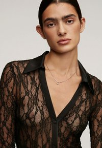 Mujer con el cabello oscuro recogido, vistiendo una blusa de encaje negro translúcido con cuello de satén y un delicado collar de plata con un pequeño colgante en forma de círculo.