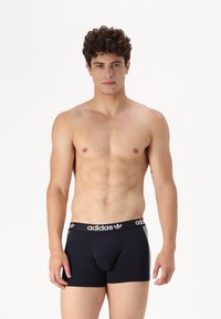 Sorte bomull boksershorts med elastisk midjebånd, prydet med adidas-logo og hvite striper på sidene. Modellen står uten skjorte.
