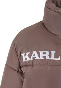 Smeđi pufasti kaput s visokim ovratnikom, s velikim bijelim logotipom "KARL" na prsima. Glatka, sjajna tkanina s kviltovanim uzorkom.
