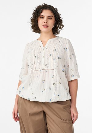 Femme aux cheveux bouclés portant un chemisier blanc à motifs floraux avec des détails en dentelle et un pantalon marron, debout sur un fond blanc uni.
