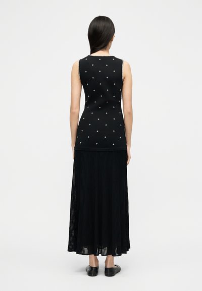 Γυναίκα που στέκεται με την πλάτη στην κάμερα, φοράει ένα αμάνικο μαύρο τοπ με μικρούς λευκούς σταυρούς και μια μακριά μαύρη πλισέ φούστα.