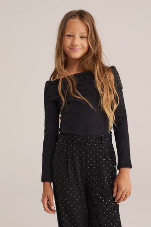 Haut noir à côtes, sans épaules, associé à un pantalon noir taille haute avec des pois blancs. Manches longues, texture lisse, coupe ajustée.