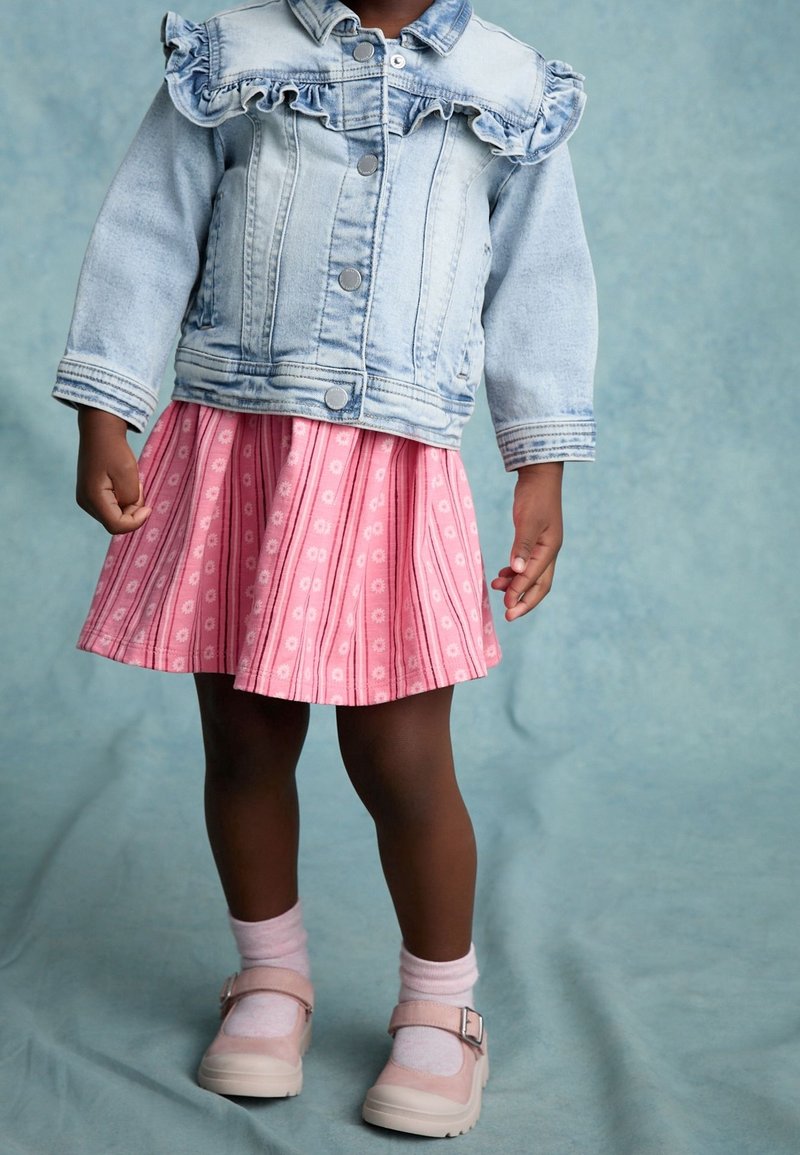 Enfant portant une veste en jean bleu clair avec détail volanté, jupe rose à motifs, chaussettes roses et chaussures roses, debout devant un fond bleu.