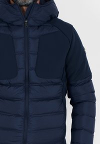 Giacca puffer blu navy con cappuccio, caratterizzata da una texture trapuntata, pannelli laterali lisci e chiusura con zip. I dettagli includono cuciture a contrasto e tasche.