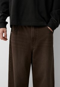 Pantalones de mezclilla marrones con un corte relajado, que cuentan con bolsillos frontales, detalles de costura sutiles y una textura suave. Combinados con una blusa negra de manga larga.