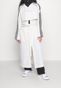 Lätt, krämfärgad trenchcoat med en kort svart huvad topp, långa ärmar och svarta ränder. Inkluderar ett skärp i midjan och sidofickor.