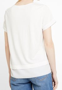 Mujer que lleva una blusa blanca de manga corta con capas y puños remangados, y jeans azules de mezclilla, vista desde atrás sobre un fondo blanco.