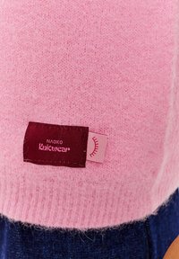 Svetloružový pletený sveter s jemnou textúrou, s tmavooranžovou etiketu, na ktorej je napísané "NAOKO Knitwear" a malým ružovým akcentom.