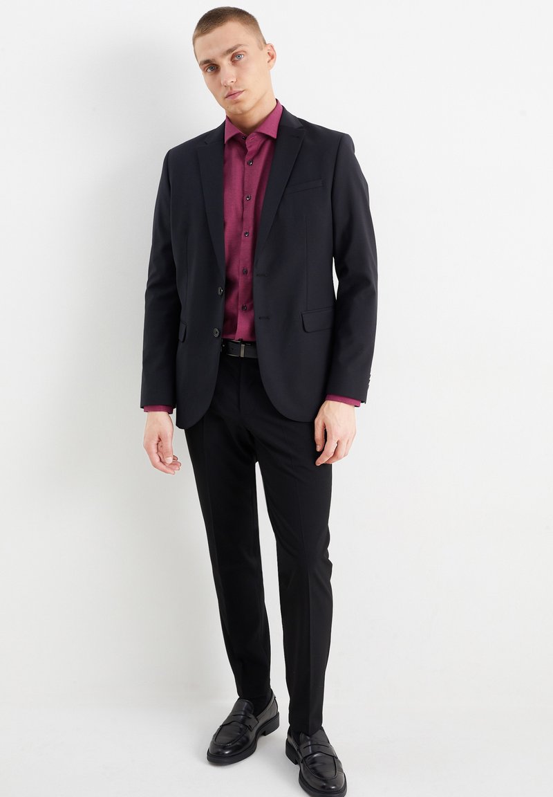 Zwarte kostuum met een getailleerd ontwerp, gecombineerd met een bordeauxkleurig button-up shirt. Glad materiaal, slanke broeken en gepolijste zwarte instappers.