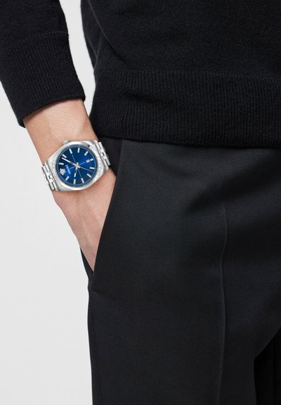 Pols met zilveren metalen horloge met een blauwe wijzerplaat, gedragen over een zwarte broek en een zwarte trui met lange mouwen, hand in de zak.