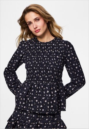 Vrouw met blond haar die een zwarte, met bloemen bedrukt, getailleerde blouse met lange mouwen en bijpassende rok draagt, staand met haar handen in de zij.