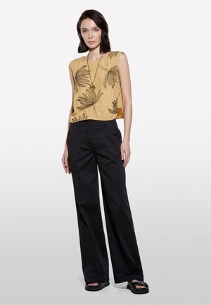 Top sans manches de couleur moutarde avec imprimé feuille noire, associé à un pantalon large noir et des sandales noires avec une boucle.