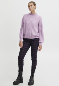 Maglione lilla a coste con spalle scese, abbinato a pantaloni a quadri neri e viola. Stivali alla caviglia neri con lacci completano il look.