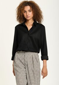 Black, long-sleeve button-up shirt with a collar, made of smooth fabric. Paired with check-patterned, high-waisted trousers.