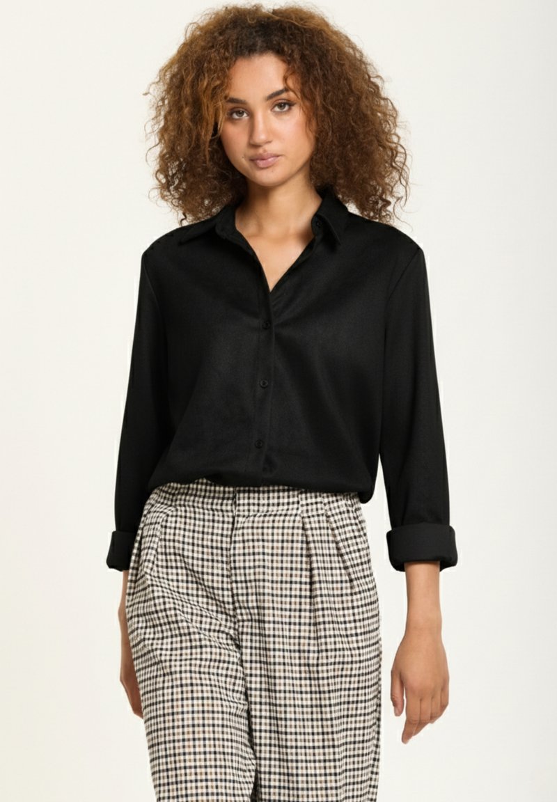 Black, long-sleeve button-up shirt with a collar, made of smooth fabric. Paired with check-patterned, high-waisted trousers.