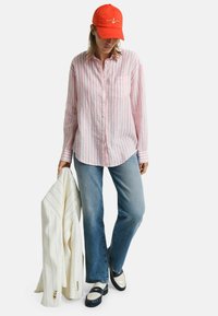 Cappellino da baseball rosso, camicia a maniche lunghe a righe rosa e bianche, jeans azzurri chiari e blazer bianco con righe, abbinati a scarpe in bianco e nero.