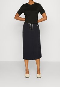 Donna che indossa una maglietta nera a maniche corte, una gonna midi blu navy con coulisse e logo GAP, e scarpe con tacco beige, in piedi con le mani dietro la schiena.
