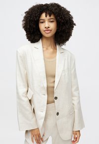 Helle beige maßgeschneiderte Blazer mit lässigem Schnitt, Ein-Knopf-Verschluss und dezenter Textur. Wird über einem beigen gerippten Top getragen, das das Outfit ergänzt.