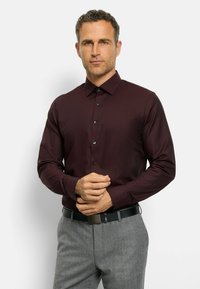 Langarmhemd in Burgunderrot mit einem subtilen Texturmuster, Button-Down-Kragen und schwarzen Knöpfen, kombiniert mit grauen Hosen und einem schwarzen Gürtel.