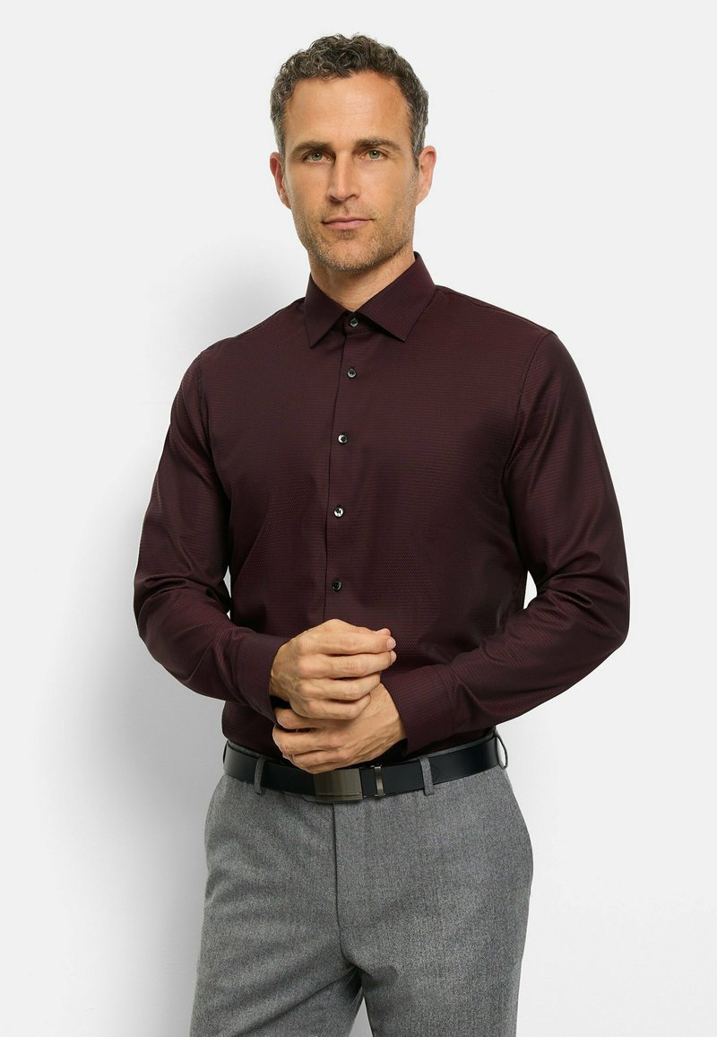 Langarmhemd in Burgunderrot mit einem subtilen Texturmuster, Button-Down-Kragen und schwarzen Knöpfen, kombiniert mit grauen Hosen und einem schwarzen Gürtel.