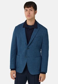 Niebieska marynarka z teksturowanej tkaniny, z dwoma przednimi kieszeniami i pojedynczym zapięciem na guzik, noszona na ciemnej koszulce polo. Dopasowany krój.