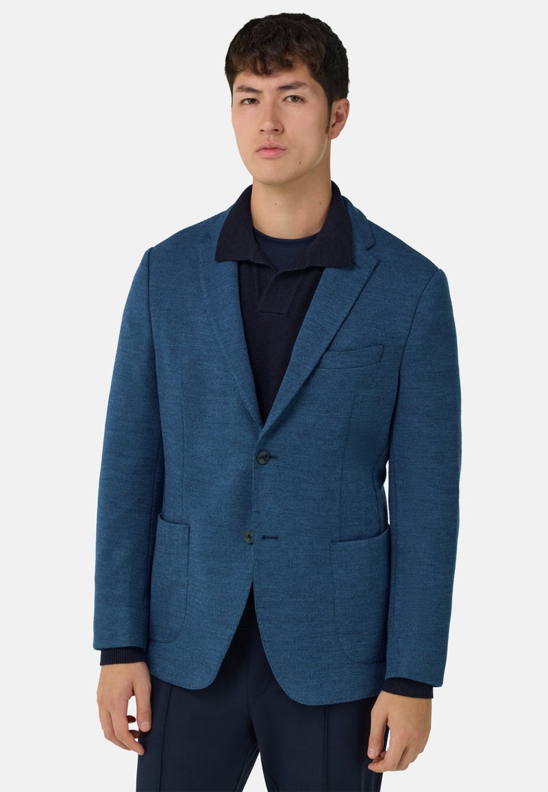 Niebieska marynarka z teksturowanej tkaniny, z dwoma przednimi kieszeniami i pojedynczym zapięciem na guzik, noszona na ciemnej koszulce polo. Dopasowany krój.