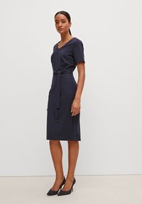 Navyblaues Kleid mit kurzen Ärmeln, V-Ausschnitt und Taillendetail. Slim-Fit mit einem knielangen Rock. Kombiniert mit schwarzen High Heels.