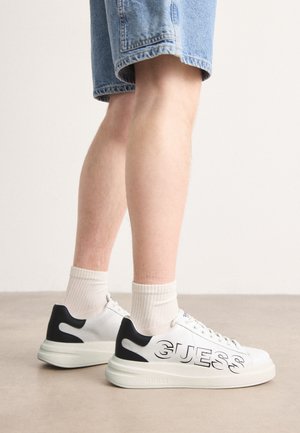 Zapatillas blancas con detalles negros, que presentan un destacado logo de "GUESS". Elaboradas con material suave, cuentan con una suela gruesa y texturizada.