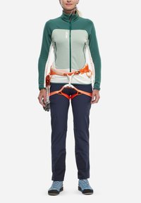Veste légère verte et blanche à zip, en tissu texturé, assortie à un pantalon bleu marine et un harnais d'escalade orange avec des boucles pour l'équipement.