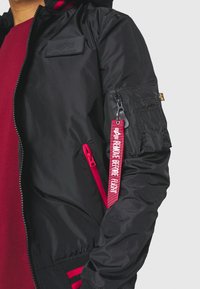 Schwarze Bomberjacke mit einer Reißverschlusstasche, rotem Akzent-Reißverschluss und einem Etikett mit der Aufschrift "VOR FLUG ENTFERNEN". Verfügt über ein strukturiertes Patch auf der Brust.