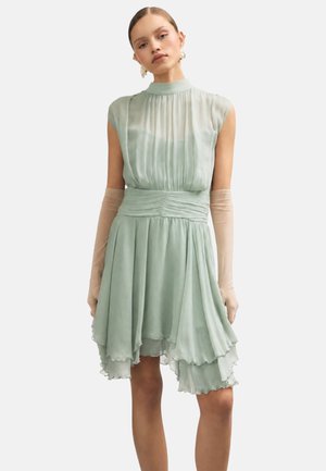 Femme portant une robe plissée vert pâle sans manches avec un ourlet à volants et des longs gants transparents beiges, debout devant un fond uni.